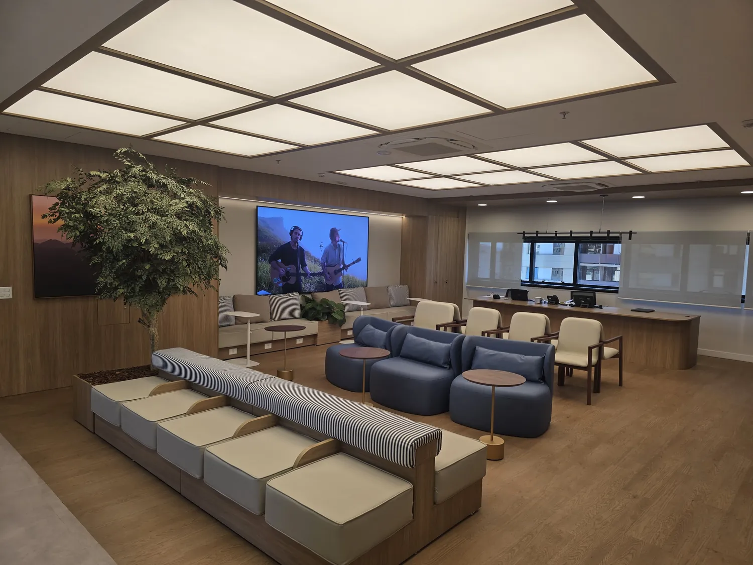 Vista diagonal do lounge: teto de lona tensionada iluminada em módulos, TV na parede e recepção ao fundo