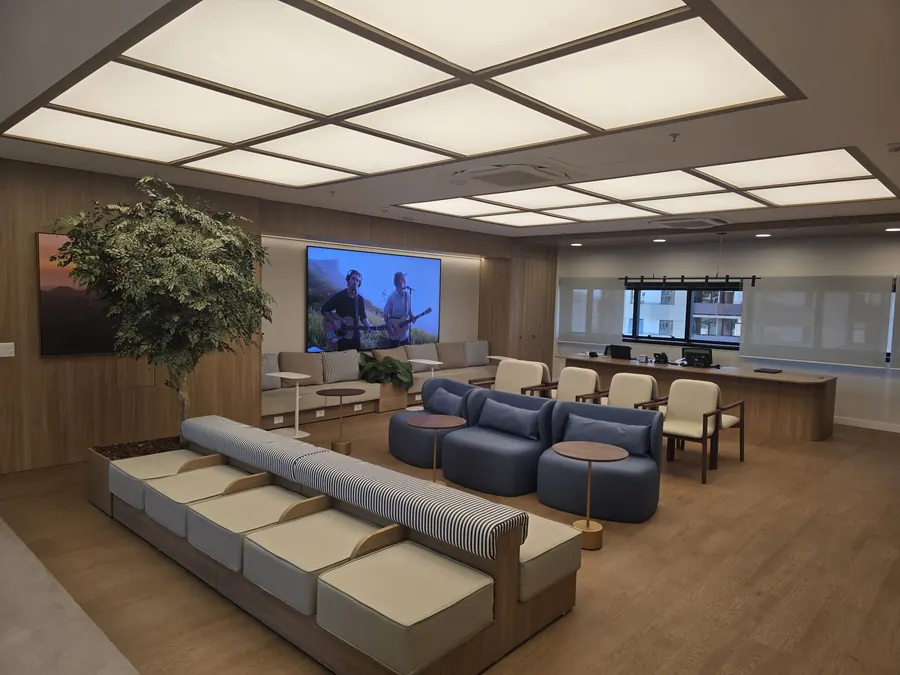 Vista diagonal do lounge: teto de lona tensionada iluminada em módulos, TV na parede e recepção ao fundo